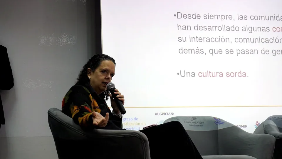 Ponente sentada en un sillón mientras habla con un micrófono frente a una pantalla de presentación durante el congreso.