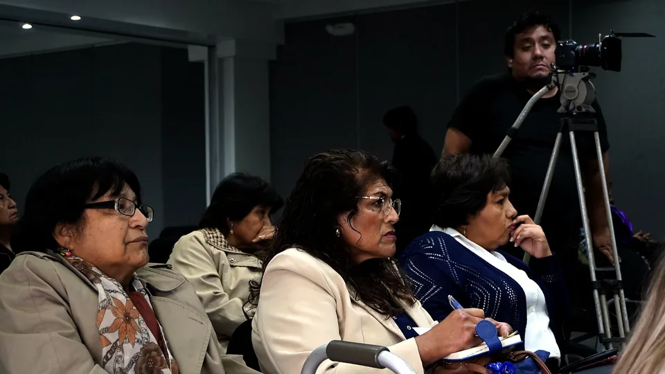 Asistentes sentadas en primera fila escuchan atentamente una ponencia; al fondo se ve una cámara en trípode.