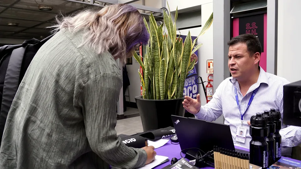 En un módulo informativo, una persona brinda orientación a una asistente frente a una mesa con materiales del evento.