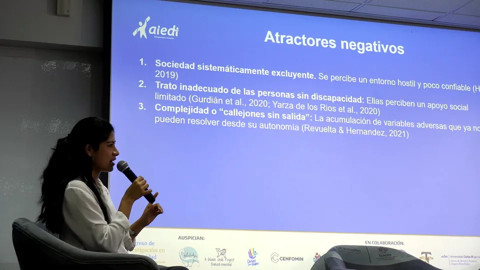Ponente sentada habla con micrófono junto a una presentación proyectada sobre barreras y discapacidad.