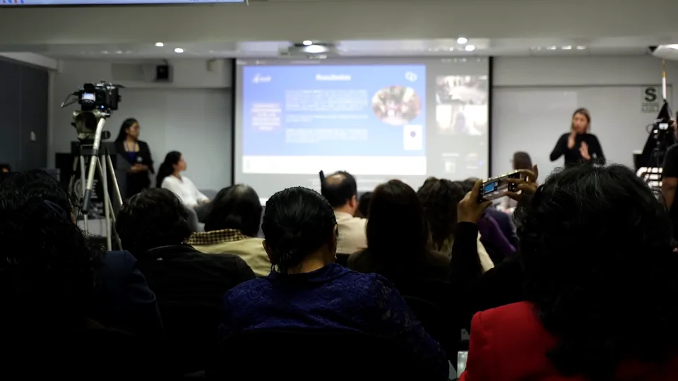 Vista general de la sala con público, cámaras y una moderadora o intérprete durante una presentación.