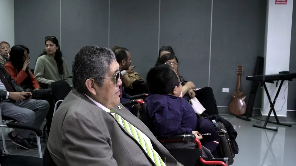 Asistentes escuchan la ponencia; en primer plano se observa a una persona con traje y otra en silla de ruedas.