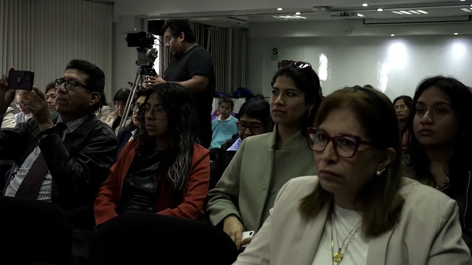 Público diverso atiende la conferencia mientras una cámara graba desde el fondo de la sala.