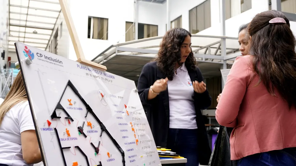 Expositora explica un plano háptico a una asistente en el área de exhibición del evento.