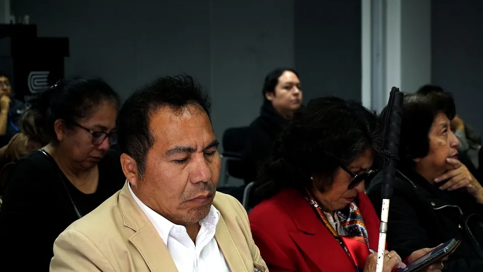 Asistentes toman notas y siguen la presentación; en primer plano se observa un bastón blanco apoyado en una silla.