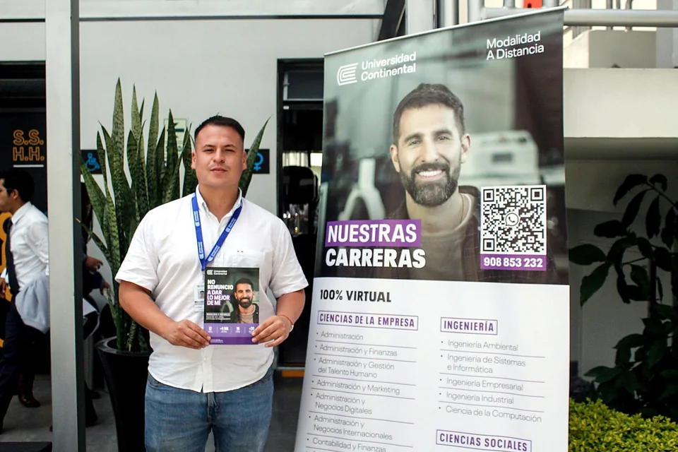 Participante posa junto a un panel promocional de la Universidad Continental sobre carreras 100 % virtuales, sosteniendo material informativo.