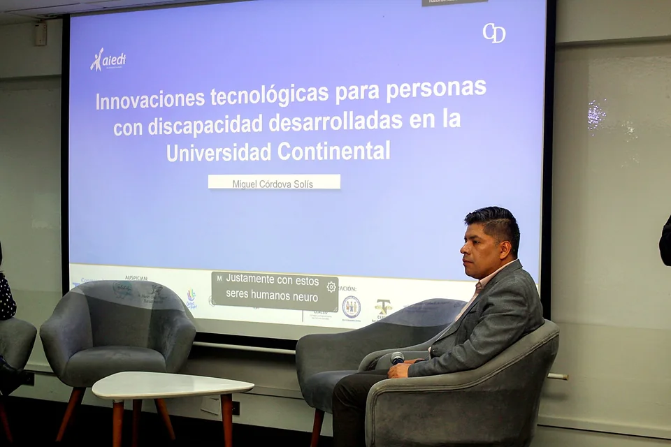 Un expositor se encuentra sentado en el escenario frente a una diapositiva titulada sobre innovaciones tecnológicas para personas con discapacidad.