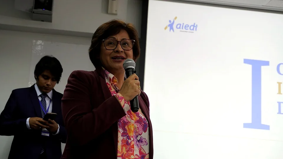 Una expositora ofrece unas palabras de bienvenida sosteniendo un micrófono frente a la pantalla principal del evento.