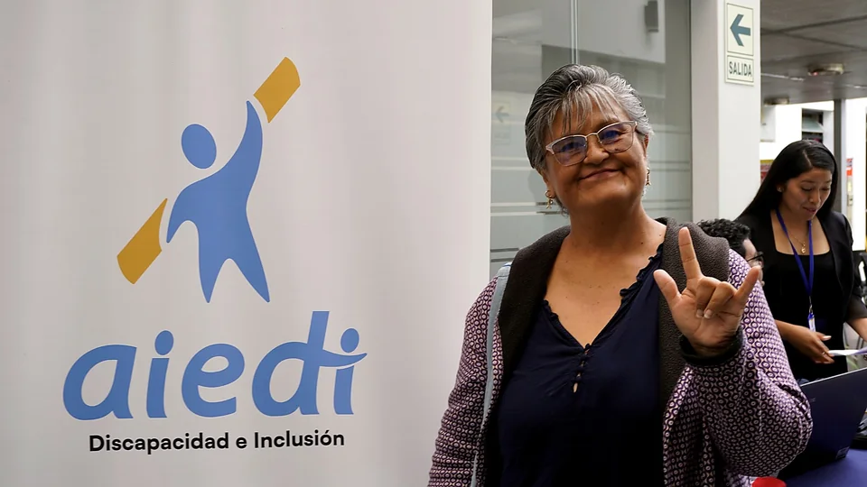 Una asistente posa sonriendo junto a un panel institucional de AIEDI y realiza una seña con la mano.