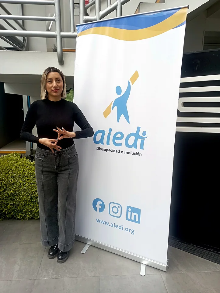 Una participante posa junto a un roll-up de AIEDI con el logotipo de la organización y sus redes sociales.