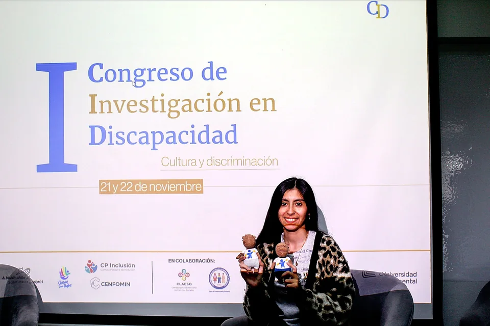 Una participante sostiene dos muñecos institucionales de AIEDI mientras posa frente a la pantalla del congreso.