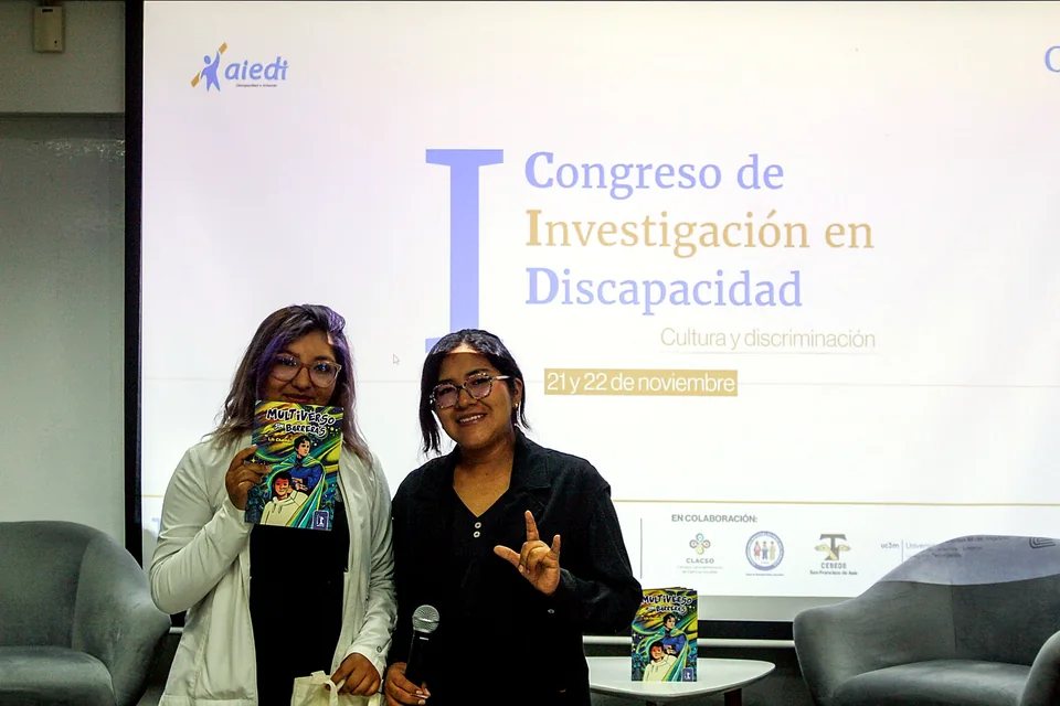 Dos participantes presentan un ejemplar del libro “Multiverso sin barreras” y posan frente a la pantalla del congreso.