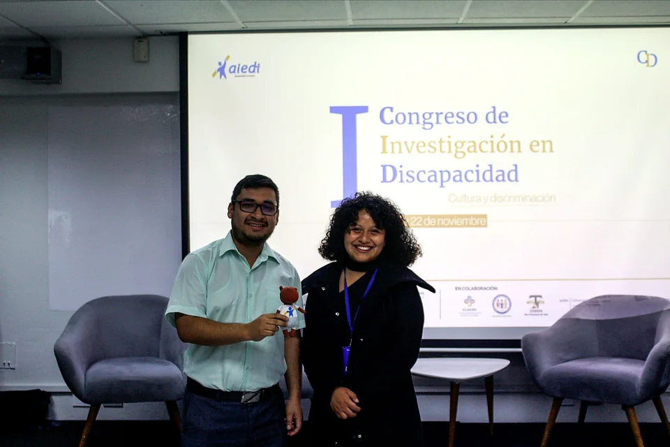 Dos participantes posan sonrientes mientras sostienen un recuerdo institucional frente al escenario del evento.