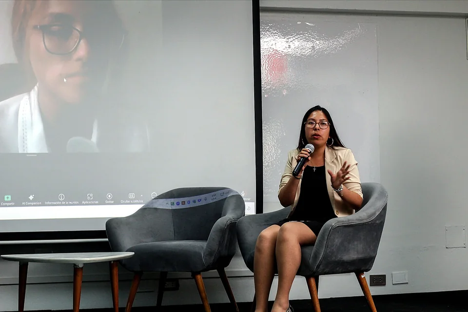 Una expositora habla con micrófono desde el escenario mientras en la pantalla se ve la participación virtual de otra persona.