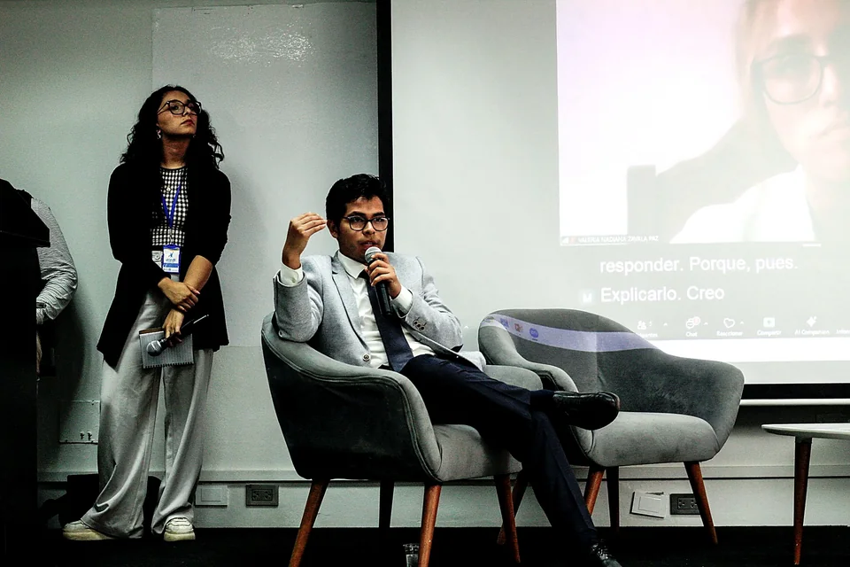 Un panelista interviene sentado en el escenario con micrófono, acompañado por una moderadora de pie y por participación remota en pantalla.